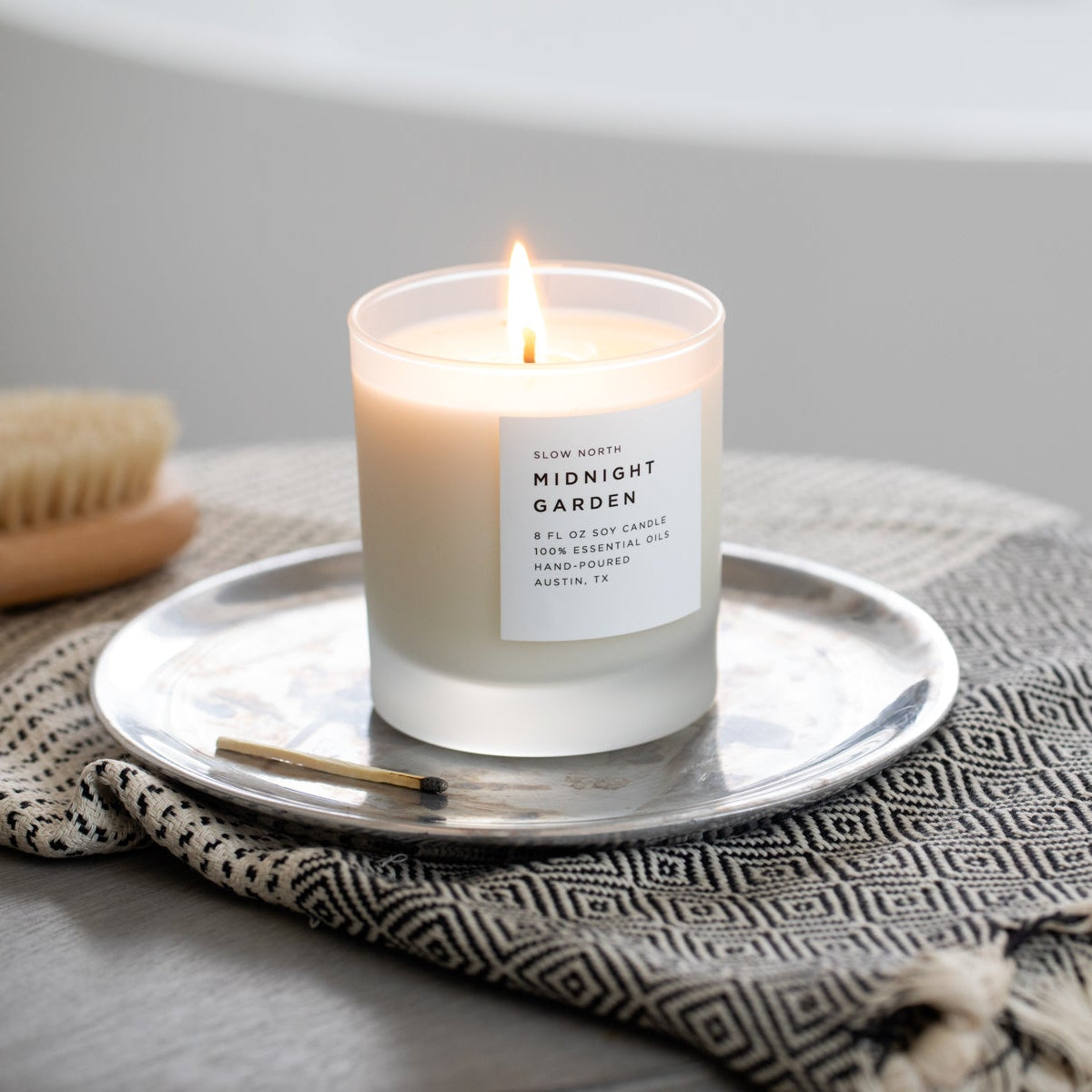 Candle labeled 'Midnight Garden' on a silver tray with a textured blanket and brush in the background.