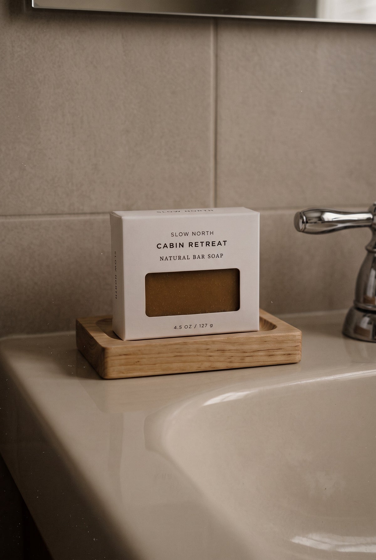 A boxed bar of soap labeled 'Cabin Retreat Natural Bar Soap' sits on a small wooden soap dish on a bathroom counter, with a sink and tiled wall in the background.