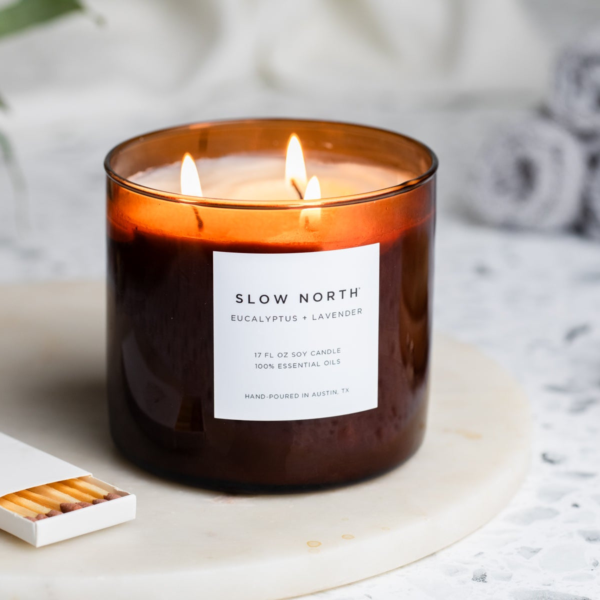 Candle with a label on a white surface with towels and a brush in the background