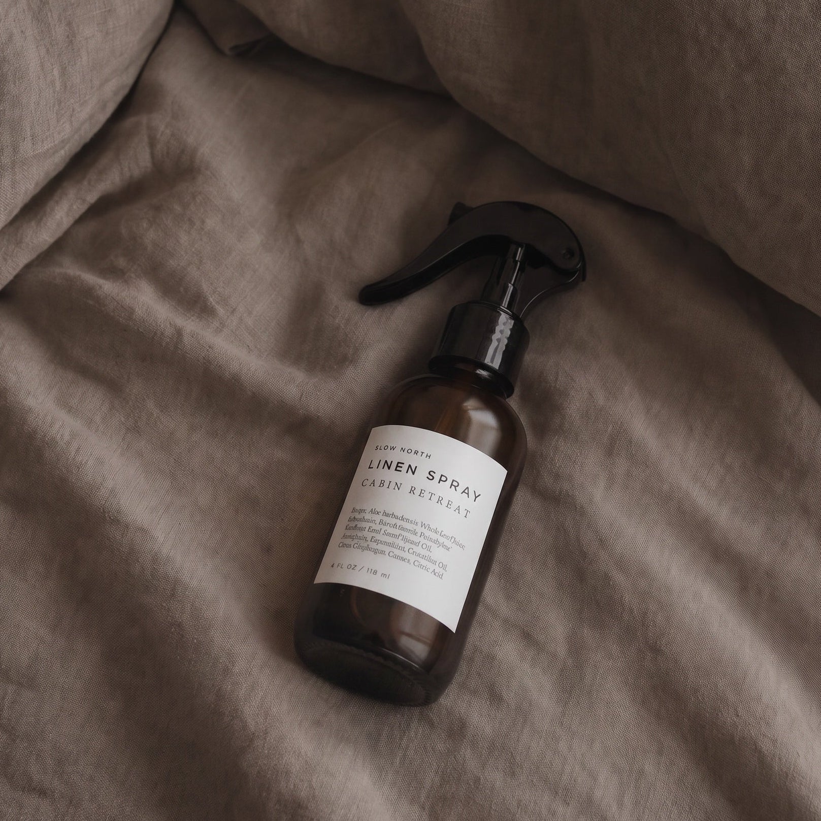 A linen spray bottle with a black spray top and white label, resting on draped brown linen fabric.